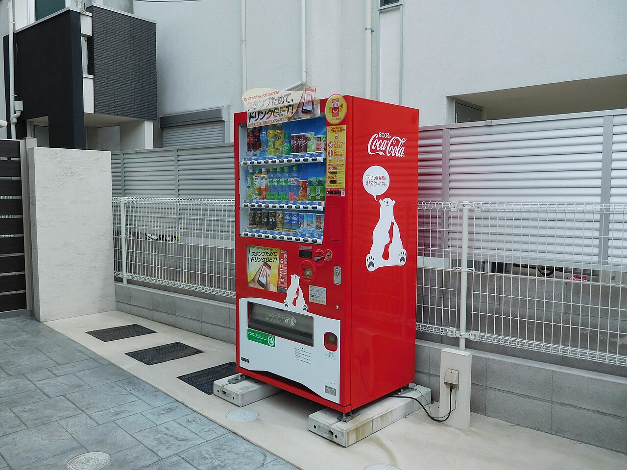 A red drink vending machine on a Tokyo street, showing rows of hot and cold beverages, a card reader, and a coin slot