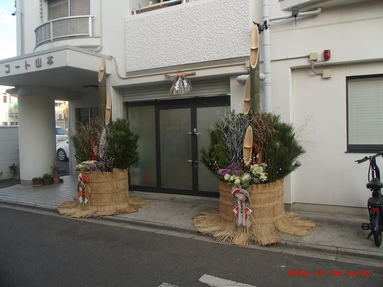 A pair of kadomatsu with diagonally-cut bamboo and pine, flanking a building entrance, with a shimenawa hung above the door